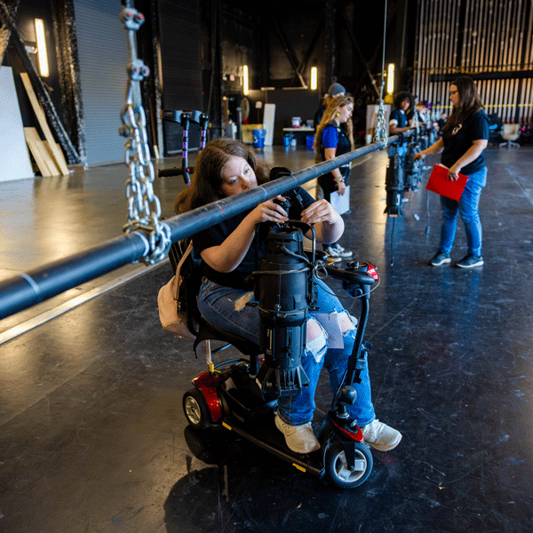 A young woman in a mobility scooter is adjusting a stage lighting fixture in a large theater space. She is wearing a black t-shirt, ripped jeans, and sneakers, with a pink backpack hanging from her seat. Several other people are working on lighting equipment in the background, including a woman holding a red clipboard. The setting is a backstage or technical training workshop, with rigging, stage lights, and various equipment visible. The space is illuminated by overhead lights, casting a professional and focused atmosphere.