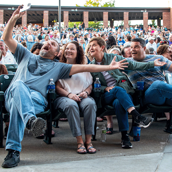 Four people are sitting in the front row of Starlight Theatre, taking a group selfie while smiling and gesturing excitedly. The stands behind them are filled with a large, diverse crowd.