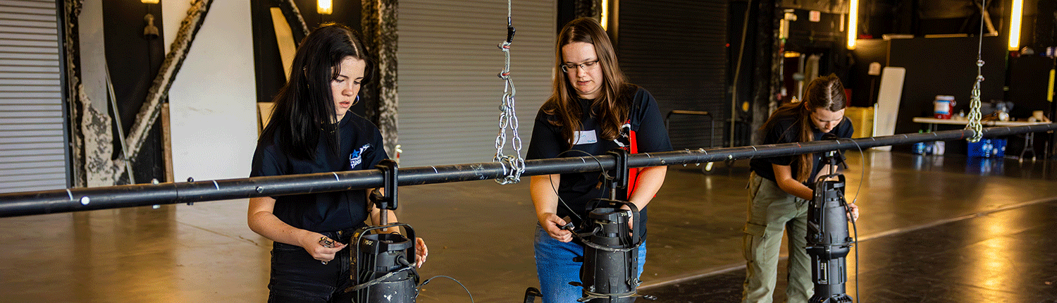 Two white girls and a white woman are working on lighting for the theatre. They are using a variety of equipment to create different lighting effects.