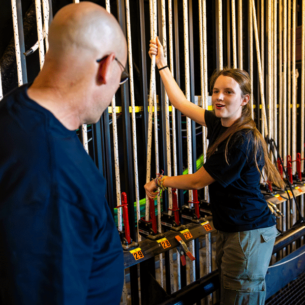 An older white male in a blue t-shirt shows a young white girl how to use pulleys for the stage.