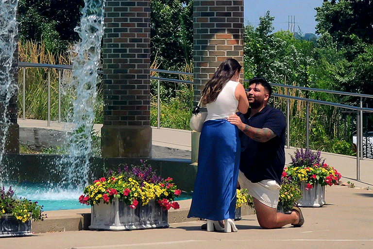 A man on his knee proposing to a woman as they stand in front of the fountain.