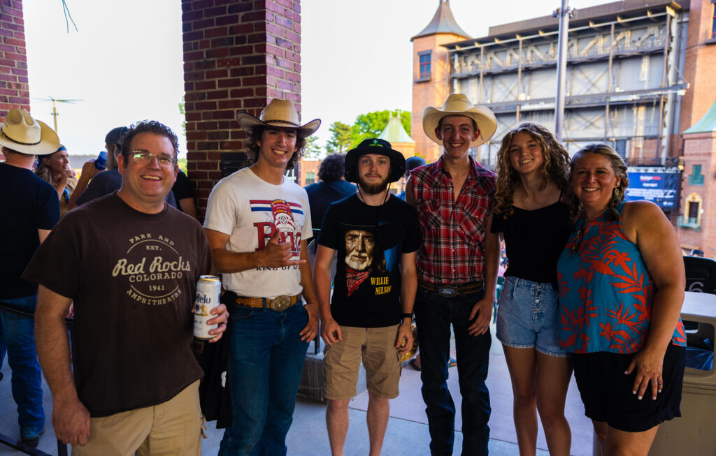 A group of smiling people posing together at an outdoor event, some holding drinks, with the Starlight Theatre in the background. An audience and stage set for live music are visible too.