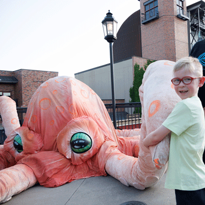 Young blonde boy wearing glasses and a light green shirt holds a tentacle of a large octopus puppet.