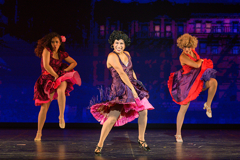 Three women wearing colorful dresses with ruffles and dancing on stage during West Side Story.